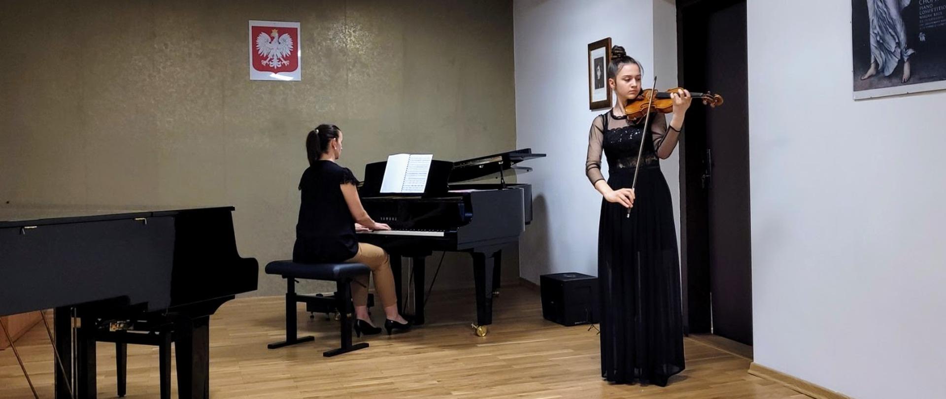Po lewej stronie zdjęcia widać akompaniatorkę grającą na fortepianie. Po prawej widać dziewczynę w czarnej sukience grającą na skrzypcach. Sytuacja ma miejsce w sali koncertowej szkoły.