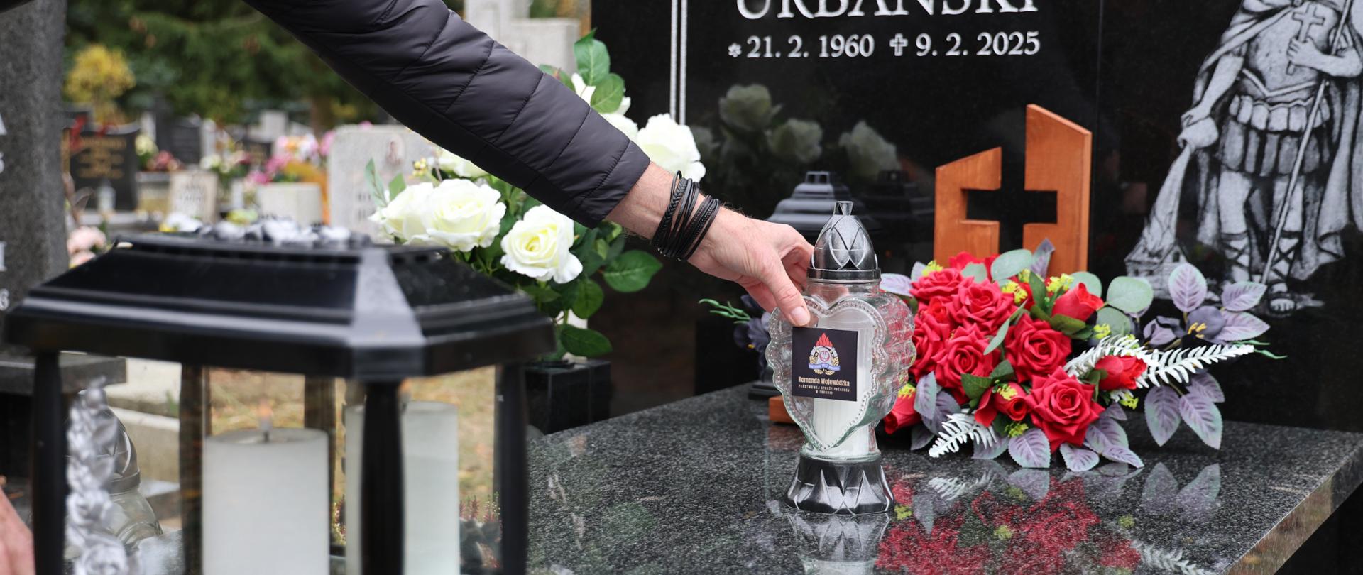 Widok na grób, pomnik na cmentarzu oraz rękę, która stawia znicz na środku płyty. Obok wiązanka czerwonych róż, Na płycie pionowej wizerunek świętego Floriana.