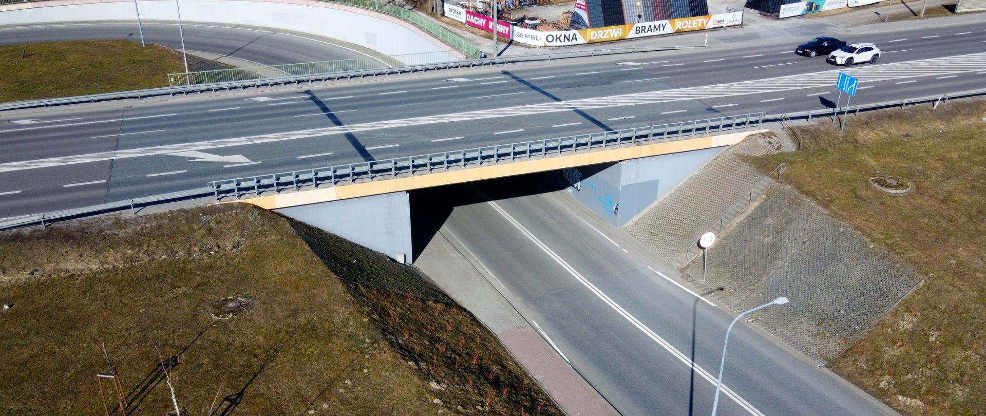 Widok z góry na drogę krajowa numer 94 w miejscowości Krasne. Jest wiosna, świeci słońce, zazieleniają się trawniki. Widać drogę z dwoma pasami w każdym kierunku, po której jadą dwa auta. Jedno auto białe, drugie czarne. Pod drogą jest zlokalizowany wiadukt. 