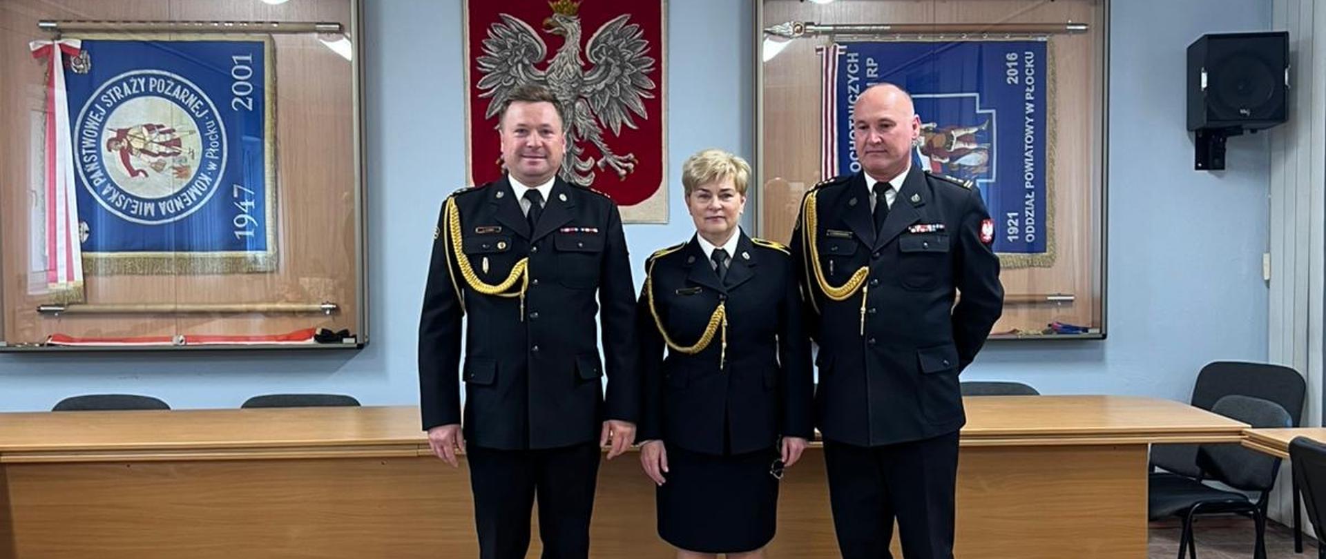 Trójka funkcjonariuszy w czarnych mundurach galowych stoi na tle godła Polski i sztandarów w sali konferencyjnej. Na podłodze przed nimi ustawiono duży kosz z czerwonymi różami przewiązanymi czerwoną wstęgą.