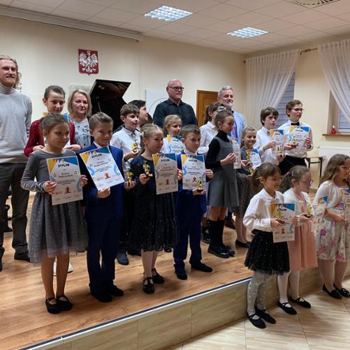 Na scenie uczestnicy III Szkolnego Konkursu "Baśniowa Karuzela" trzymają dyplomy i statuetki. Między nimi jurorzy, dyrekcja szkoły oraz organizator konkursu. Wszystkie dzieci ubrane na galowo. W tle fragment fortepianu a na ścianie godło Polski. 