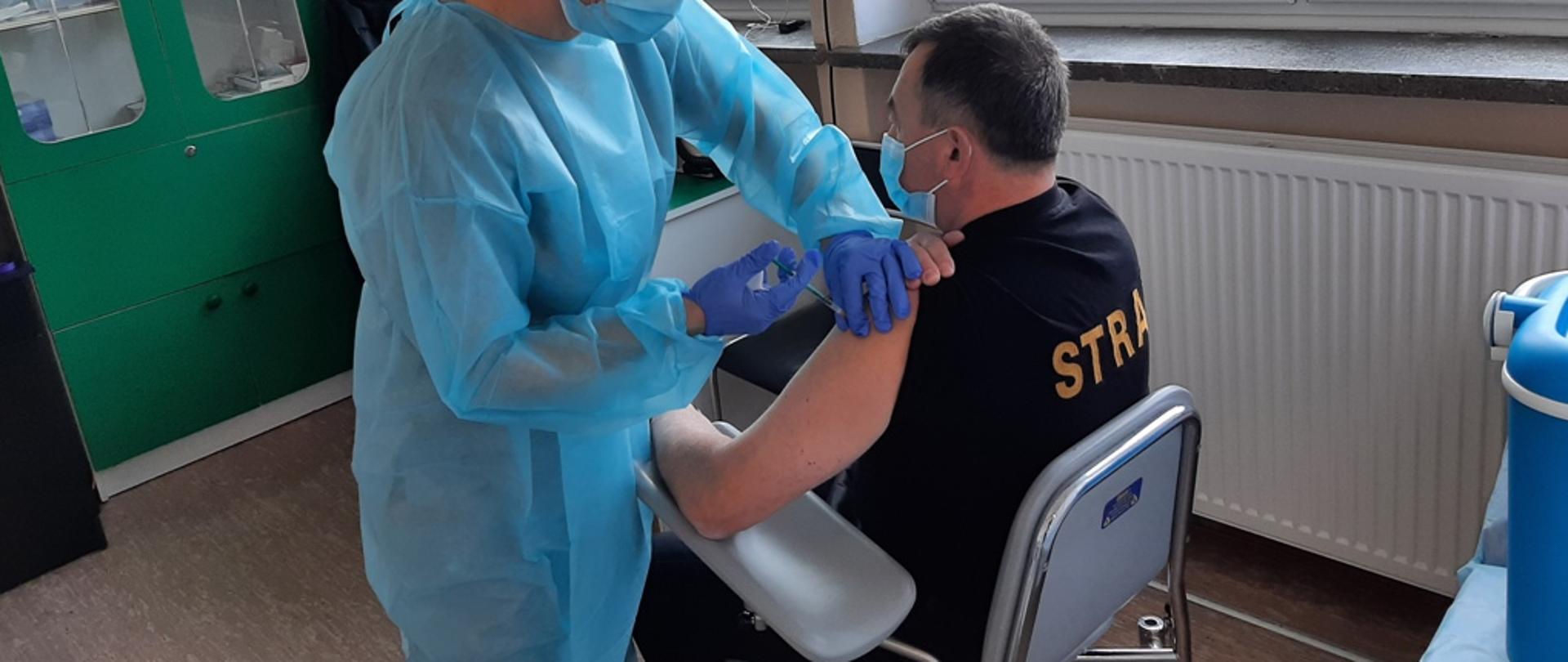 Kolorowa fotografia przedstawia na pierwszym planie strażaka w czarnej koszulce z napisem na plecach STRAŻ, siedzącego na krześle, któremu pielęgniarka podaje szczepionkę. W tle widok gabinetu szczepień.