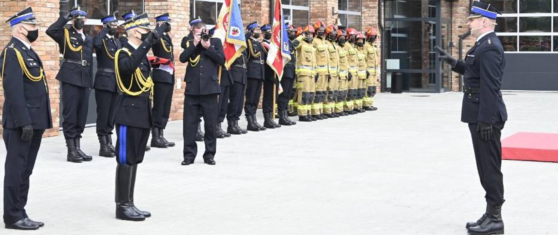 Po lewej stronie na pierwszym planie widać strażaków w umundurowaniu galowym, strażacy salutują, jeden z nich robi trzyma w dłoniach aparat i robi zdjęcie strażakowi, który stoi po lewej stronie. Na dalszym planie widać strażaków w ubraniach specjalnych a w tle budynek.