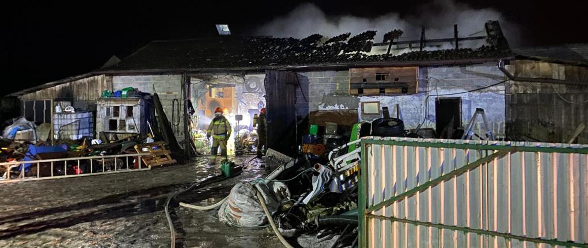 Zdjęcie przedstawia murowany garaż. Z dachu garażu wydobywa się biały dym. Z wnętrza widać sylwetki dwóch strażaków w umundurowaniu specjalnym oraz w aparatach ochrony dróg oddechowych. Przed budynkiem garażu różne narzędzie i sprzęty rolnicze. Zdjęcie zrobione w porze nocnej. 