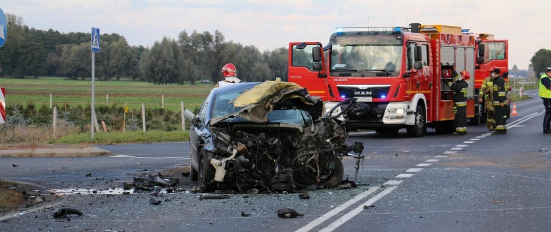 Zdjęcie przedstawia uszkodzony pojazd osobowy oraz samochody pożarnicze i strażaków. 