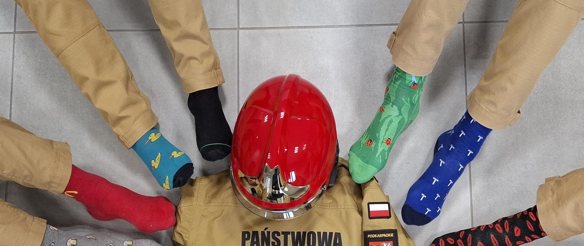 Widok z góry na okrąg utworzony przez stopy osób w nogawkach od piaskowych mundurów strażackich i kolorowych, niepasujących do siebie skarpetkach. W centrum okręgu leży kurtka mundurowa z napisem „PAŃSTWOWA STRAŻ POŻARNA” oraz czerwony hełm strażacki.