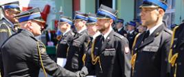 Oficer Państwowej Straży Pożarnej wręcza dyplom gratulacyjny i podaje rękę uśmiechniętemu strażakowi stojącemu w szeregu. Obok stoją inni nagrodzeni funkcjonariusze w mundurach galowych.