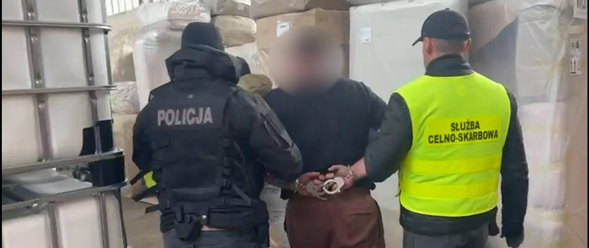 Dwóch funkcjonariuszy eskortuje osobę z rękami skutymi z tyłu przez halę magazynową, w której znajdują się duże pojemniki i opakowania.