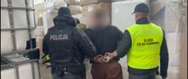 Dwóch funkcjonariuszy eskortuje osobę z rękami skutymi z tyłu przez halę magazynową, w której znajdują się duże pojemniki i opakowania.