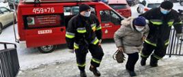 Na zdjęciu widać dwóch ratowników OSP i starsza kobietę idących po schodach do góry. Jeden ratownik trzyma kobietę w podeszłym wieku za łokieć a drugi po lewo kadru idzie obok. Kobieta trzyma w prawej dłoni małą torebkę. Ratownicy są ubrani w ubranie typu Nomex z odblaskami barwy jasnożółtej i szarej, na głowie mają czapkę wełnianą barwy czarnej, maseczkę na twarzy oraz rękawiczki jednorazowe. W tle samochody osobowe. Na ziemi widać śnieg. Auto jest typu furgon barwy czerwonej z napisem na prawych przednich drzwiach OSP Stary Waliszew, po lewej stronie auta widnieje numer operacyjny samochodu oraz napis Straż. W tle samochody osobowe.