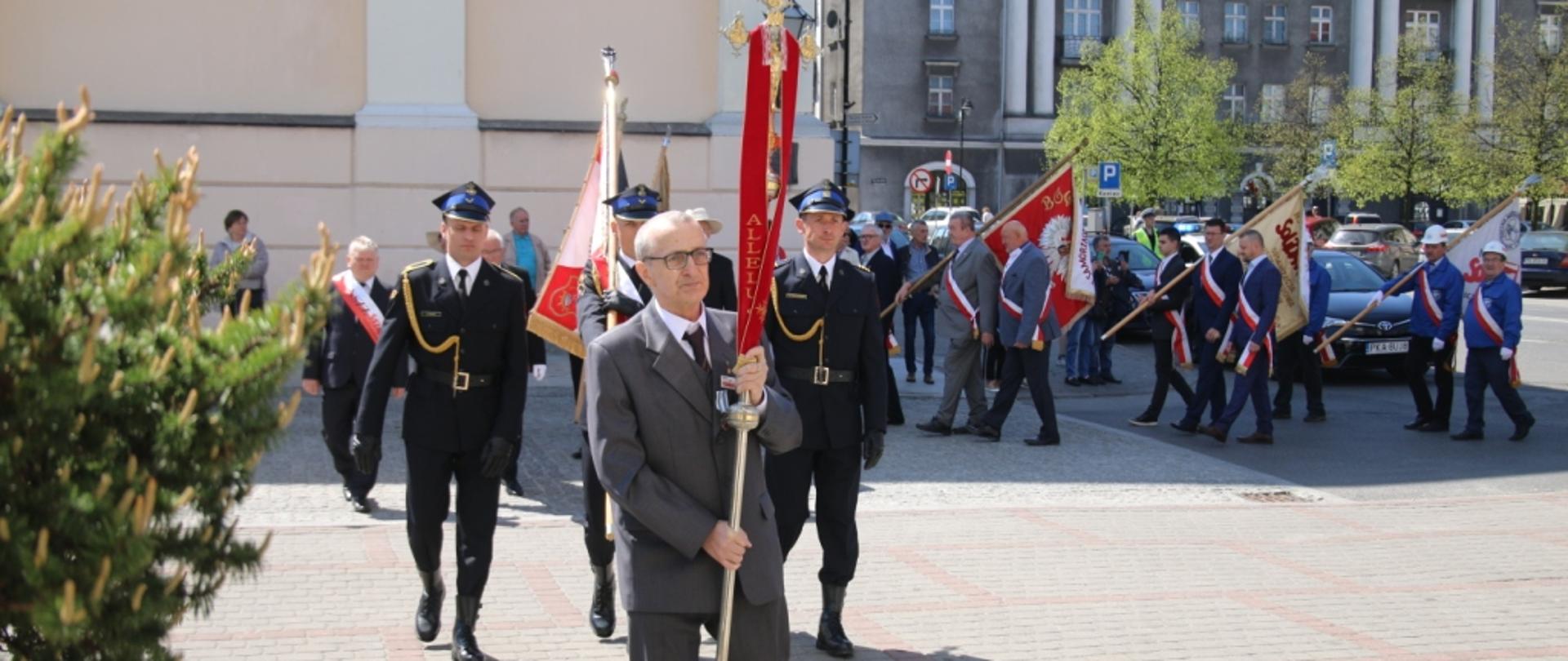 Poczty sztandarowe podczas przemarszu, na przedzie mężczyzna niosący krzyż, za nim sztandar niesiony przez poczet w mundurach wyjściowych Państwowej Straży Pożarnej
