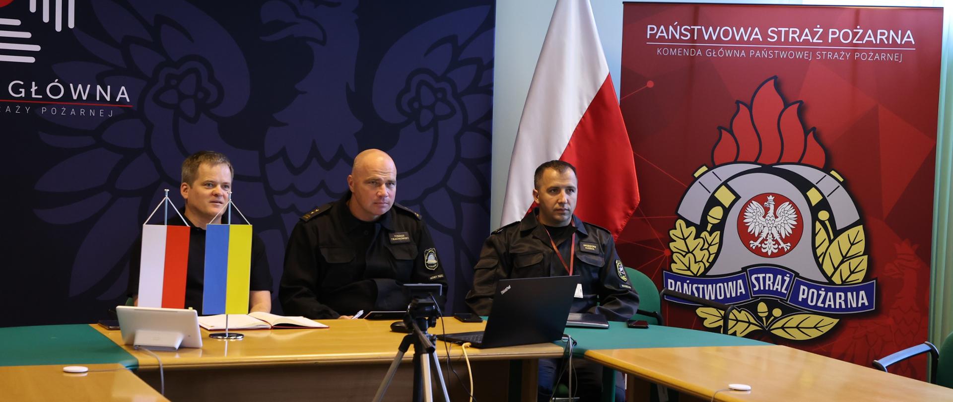 Na zdjęciu widać przedstawicieli Komendy Głównej Państwowej Straży Pożarnej siedzących przy stole konferencyjnym w czasie wideokonferencji z przedstawicielami straży pożarnej Ukrainy. W tle banery z logo PSP. 