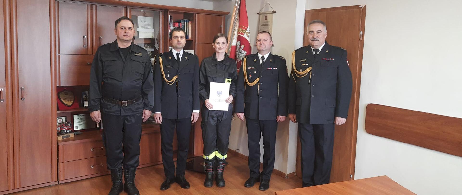 Pięcioro strażaków stojących w pomieszczeniu biurowym. Za strażakami w stojaku na podłodze po prawej stronie znajduje się sztandar .