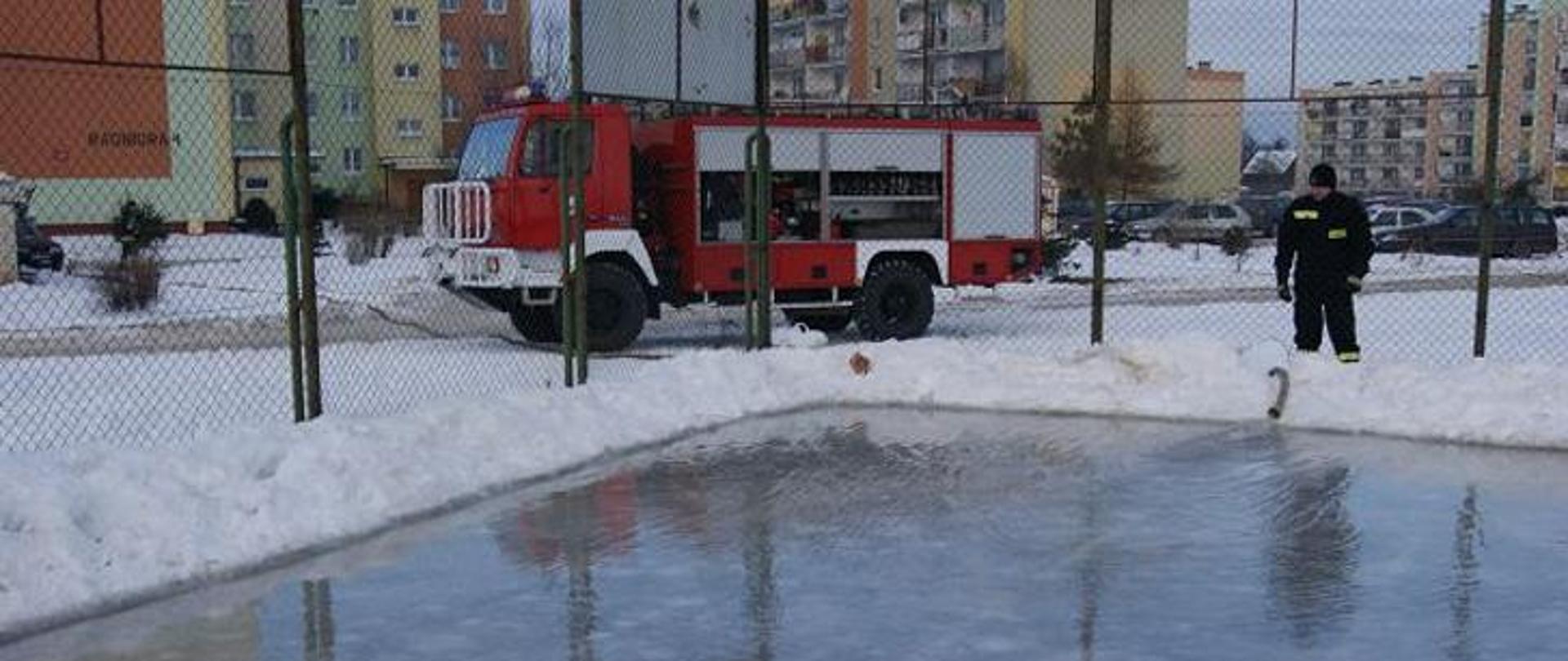 Zdjęcie przedstawia wóz strażacki oraz strażaka, który przygotowuje lodowisko na boisku szkolnym.