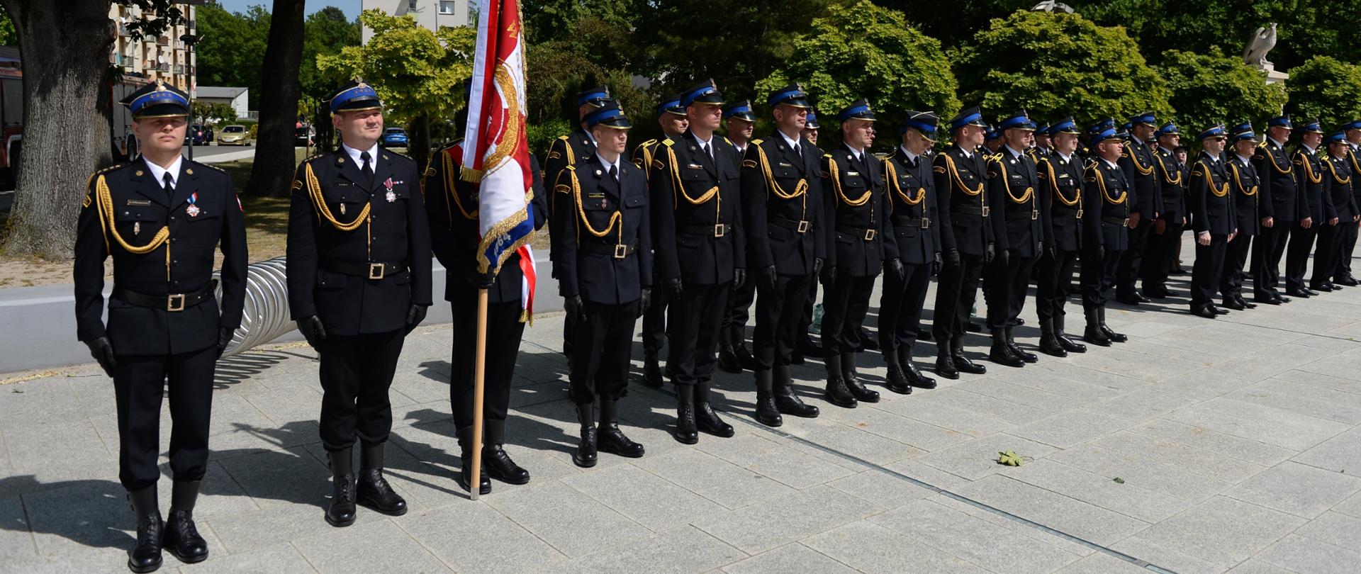 Zdjęcie zrobione na zewnątrz. Strażacy w ugrupowaniu stoją na uroczystym apelu