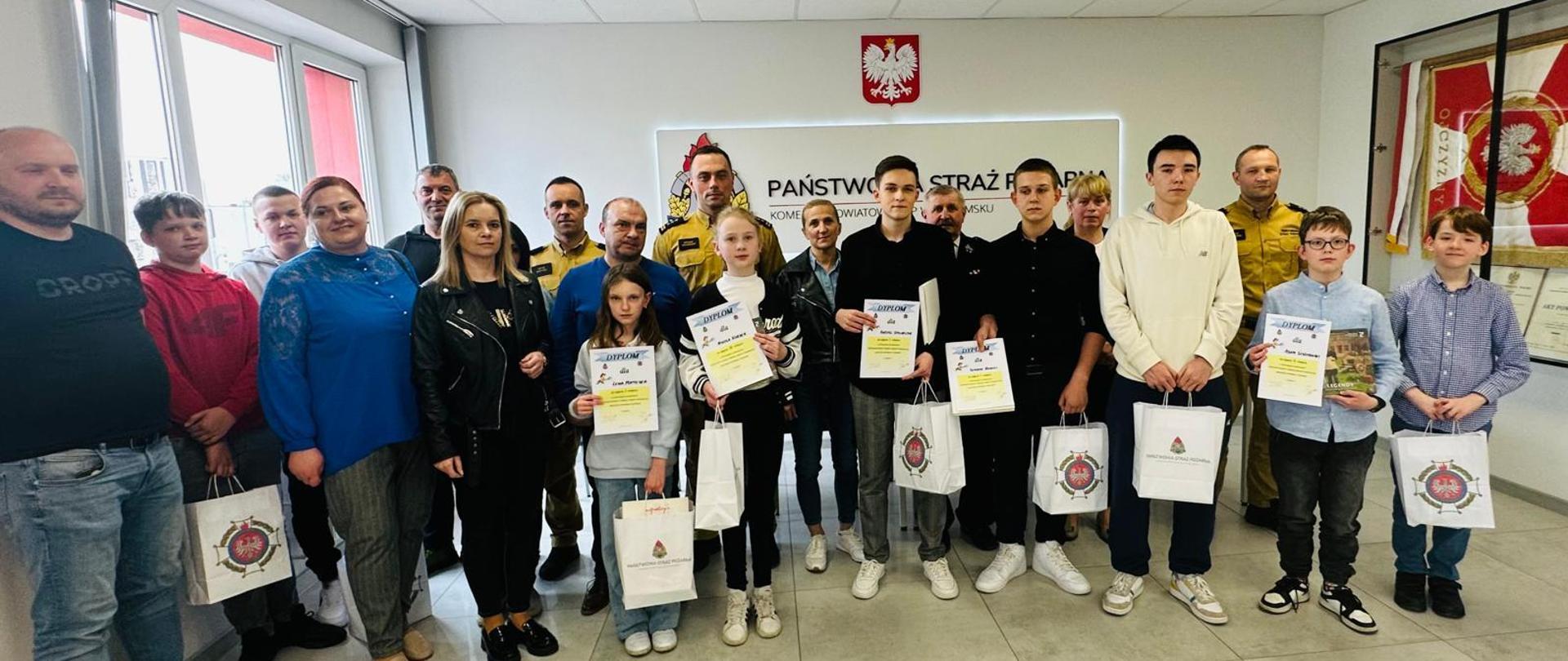 Zdjęcie grupowe na pierwszym planie funkcjonariusze – strażacy w umundurowaniu oraz finaliści konkursu – dzieci trzymające nagrody oraz dyplomy wraz w opiekunami.