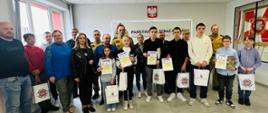 Zdjęcie grupowe na pierwszym planie funkcjonariusze – strażacy w umundurowaniu oraz finaliści konkursu – dzieci trzymające nagrody oraz dyplomy wraz w opiekunami.