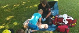 Zdjęcie przedstawia dzieci oraz strażaka siedzących na trawie. Strażak udziela informacji na temat udzielania kwalifikowanej pierwszej pomocy oraz przygląda się uczniowi, który wykonuje RKO na manekinie. 
