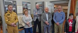 Na zdjęciu podczas wernisażu wystawy „Na STRAŻY bezpieczeństwa mieszkańców Ziemi Gorlickiej" widoczni w ubraniu służbowym koloru piaskowego strażak i w ubraniach cywilnych zaproszeni na wernisaż goście 7 osób. W tle widoczne zdjęcia i eksponaty pożarnicze. 