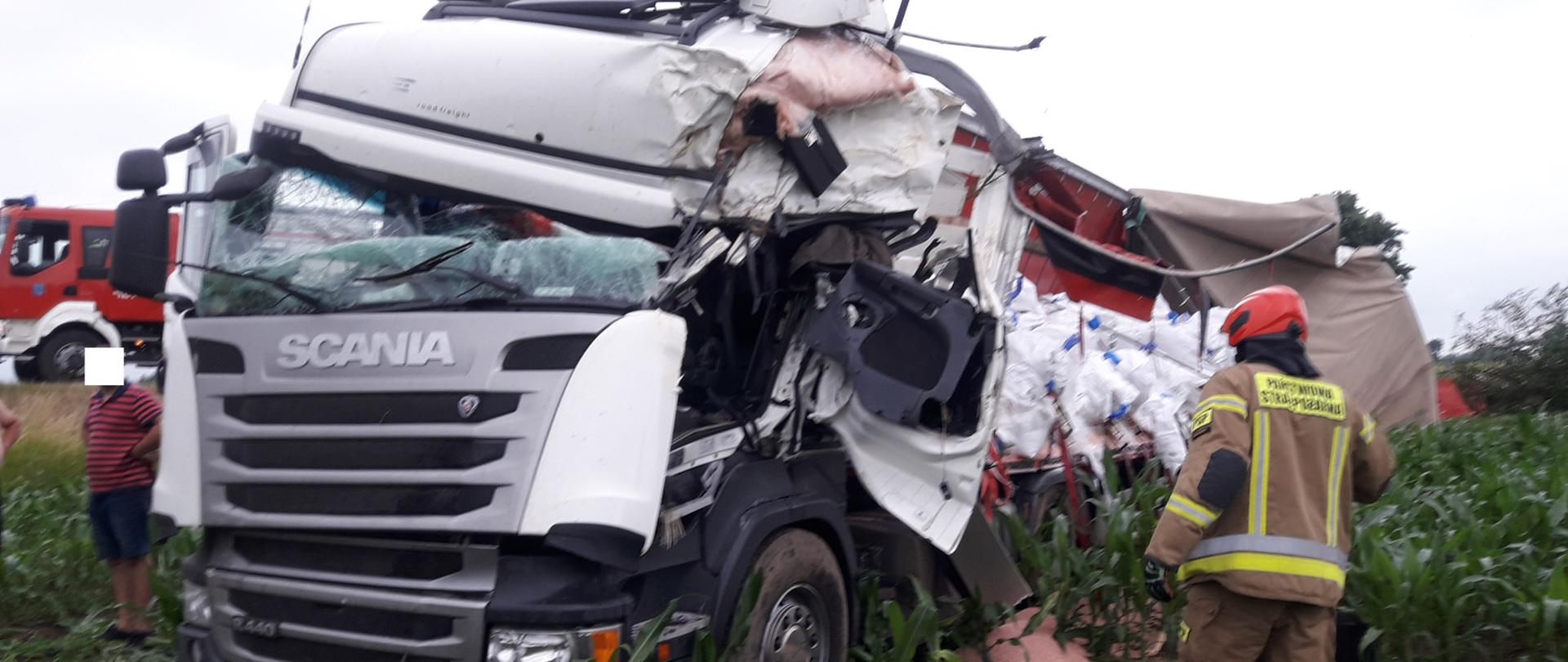 Rozbity samochód ciężarowy, przy nim strażak PSP. W tle samochód pożarniczy.