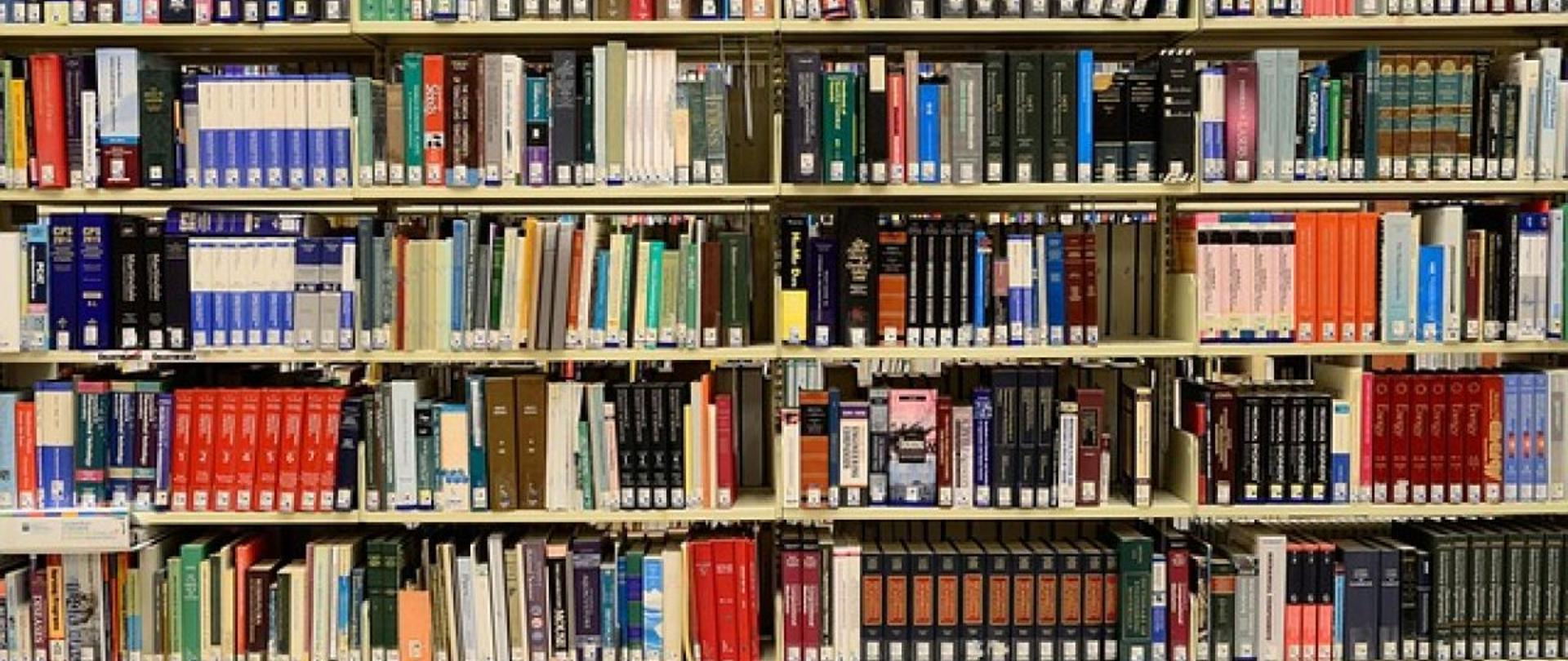Regał biblioteczny wypełniony książkami o różnych kolorach i rozmiarach. Publikacje są uporządkowane na kilku półkach i oznaczone etykietami bibliotecznymi.