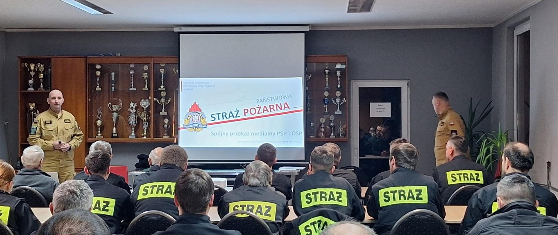 Na zdjęciu znajdują się strażacy w ubraniach koszarowych, którzy siedzą w ławkach. W tle stoi strażak, na ekranie wyświetlona jest prezentacja