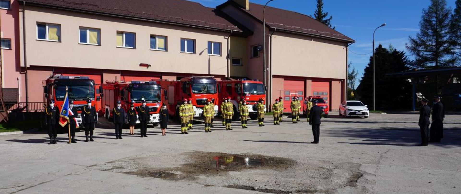 Na placu przed czerwonymi wozami bojowymi stoją strażacy w szyku ze sztandarem w mundurach granatowych oraz w ubraniach bojowych.