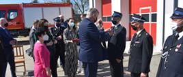 Zdjęcie przedstawia strażaków stojących w szeregu. Strażakom nadawane są odznaczenia przez inne znajdujące się na uroczystości osoby. Wśród osób wręczających odznaczenia są kobiety i mężczyźni. Strażacy ubrani są w mundury wyjściowe. Wszyscy mają na twarzy założone maseczki chirurgiczne. W tle zdjęcia widać bramy garażowe remizy OSP oraz stojący samochód pożarniczy. 