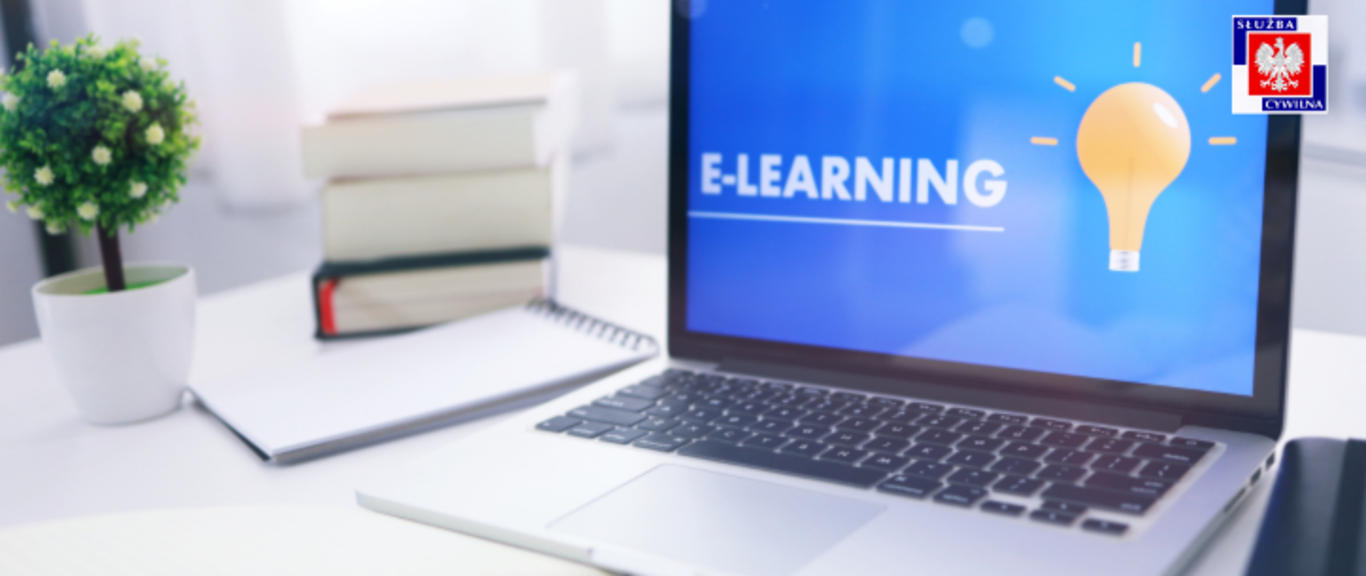 grafika do kursów e-learningowych : na biurku widać laptop (na ekranie żarówka i napis e-learning), obok stos książek i doniczka z kwiatkiem