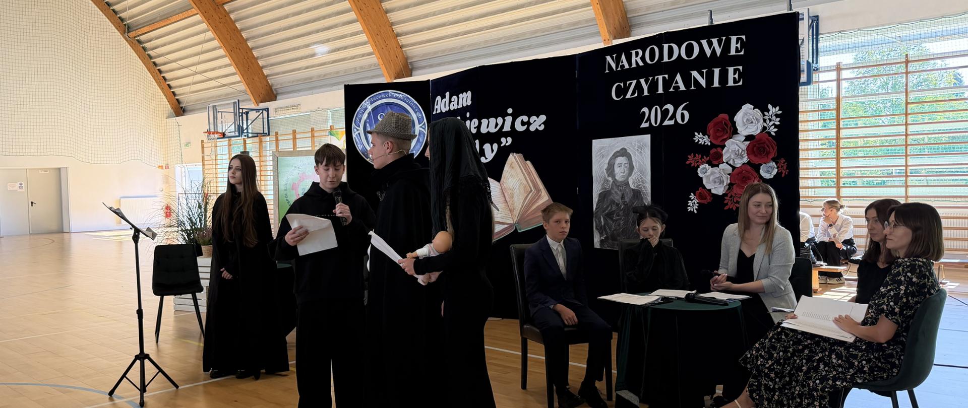 Cztery młode osoby w czarnych płaszczach czytają fragmenty utworu na scenie