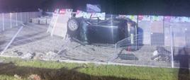 Samochód leży na boku posesji ze sprzedażą kostki brukowej. Auto wcześniej uszkodziło ogrodzenie posesji. 