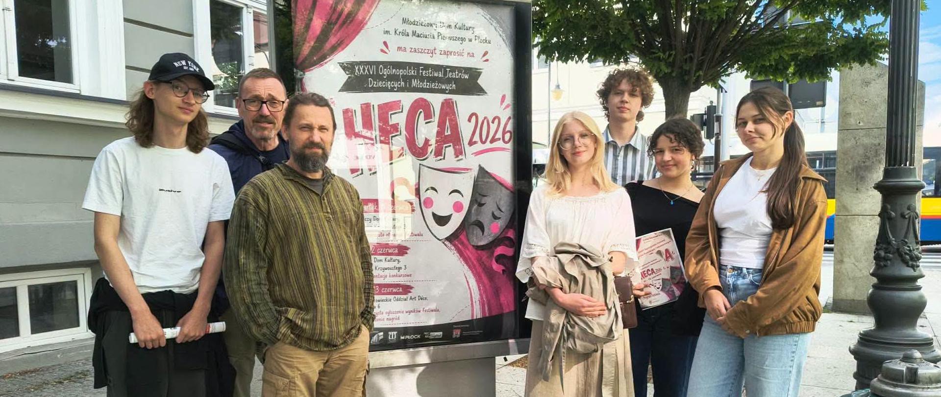 Przed budynkiem i pod drzewem jest umieszczony na betonowym stelażu duży afisz informujący o XXXVI Ogólnopolskim Festiwalu Teatrów Dziecięcych i Młodzieżowych HECA 2026. Z lewej strony stoi chłopak i dwóch mężczyzn, a z prawej strony afisza stoją trzy dziewczyny i chłopak za nimi