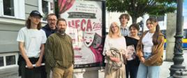 Przed budynkiem i pod drzewem jest umieszczony na betonowym stelażu duży afisz informujący o XXXVI Ogólnopolskim Festiwalu Teatrów Dziecięcych i Młodzieżowych HECA 2026. Z lewej strony stoi chłopak i dwóch mężczyzn, a z prawej strony afisza stoją trzy dziewczyny i chłopak za nimi