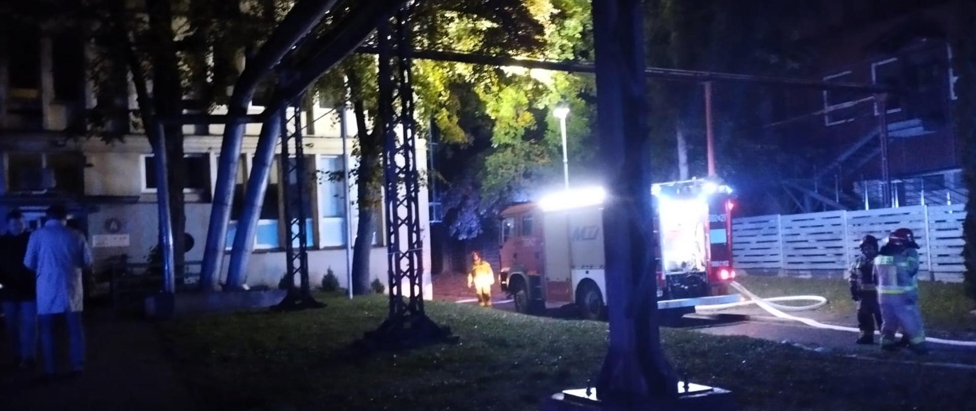 Zdjęcie przedstawia: w głębi halę przemysłową, gdzie doszło do pożaru dachu. Przy niej z prawej strony stoi samochód pożarniczy z rozciągniętymi wężami gaśniczymi, obok niego ratownicy. Widać też estakadę po której biegną linie przesyłowe centralnego ogrzewania, a z lewej strony ewakuowani pracownicy.