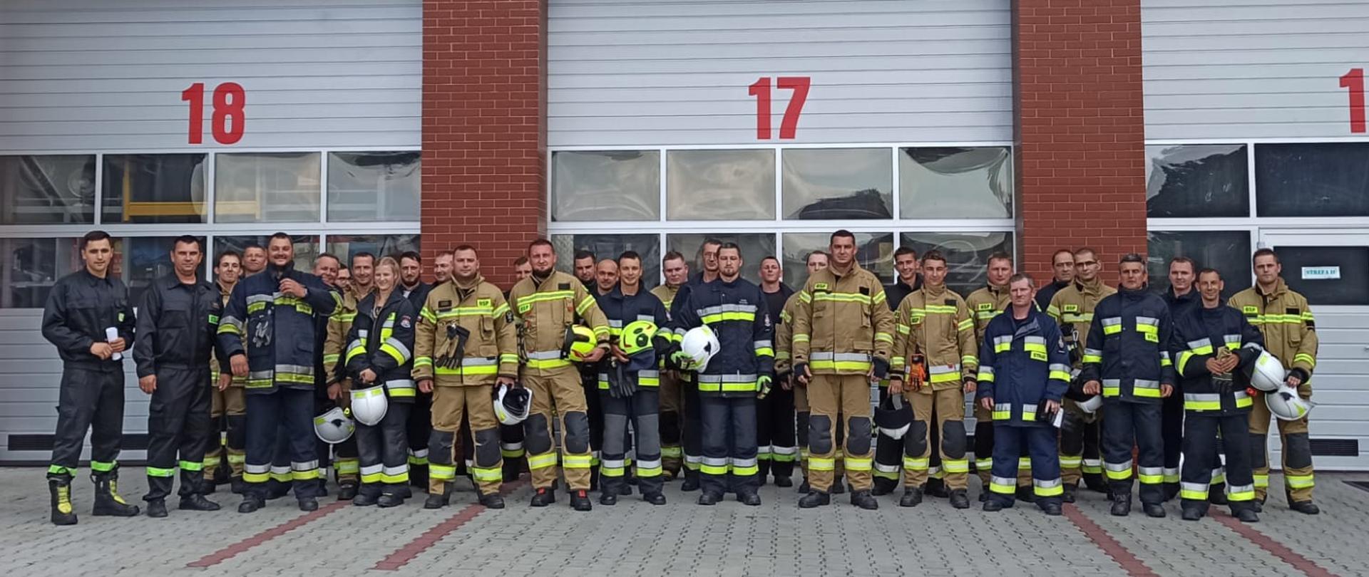 Zdjęcie ukazuje grupę strażaków, którzy przystępują do szkolenia kierowców-konserwatorów OSP. Strażacy stoją na placu ubrani w ubrania bojowe i koszarowe. W tle widać bramy garażu komendy powiatowej PSP w Bochni