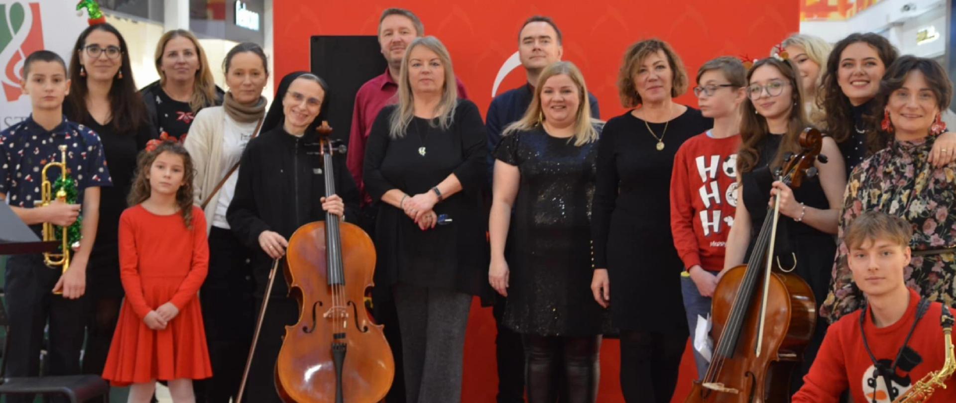 Wykonawcy koncertu na czerwonym dywanie na tle czerwonej ścianki. Niektórzy trzymają instrumenty o skrzypce, trąbka, wiolonczela, saksofon