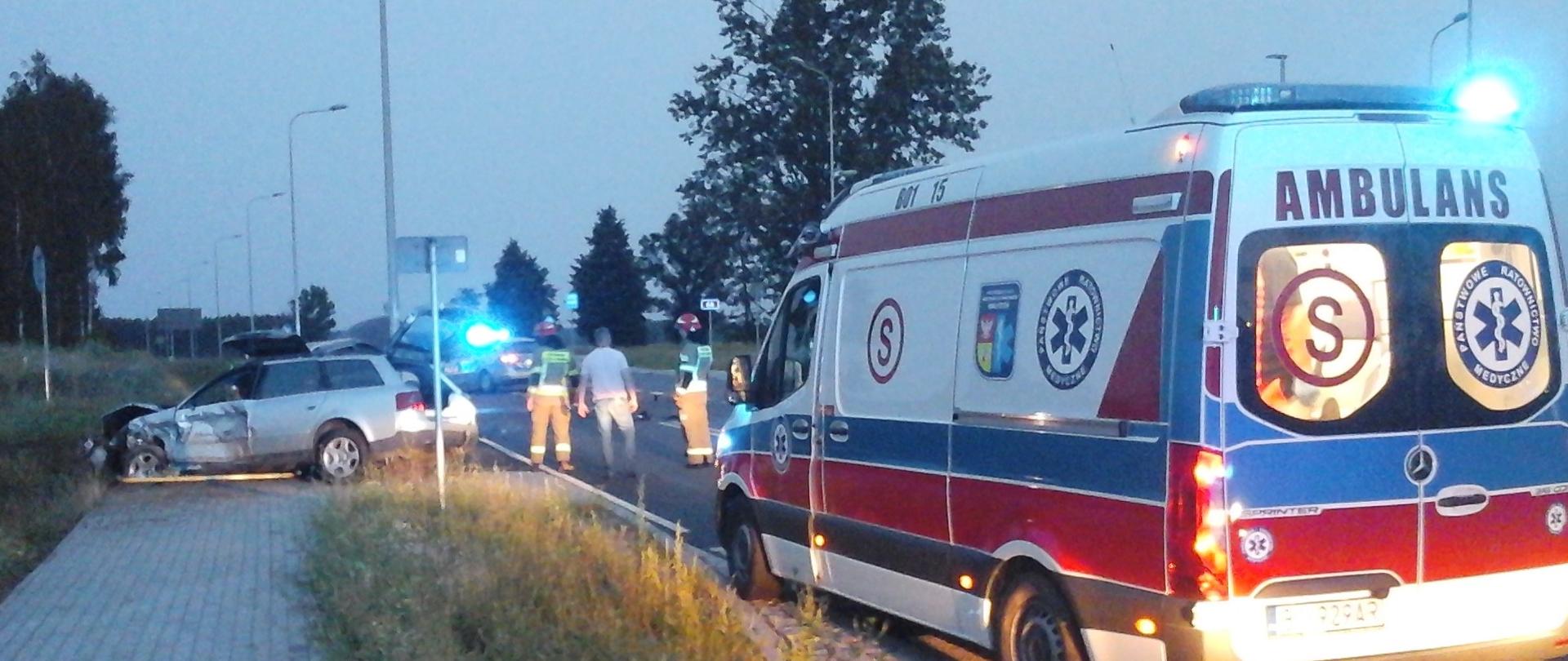 Zespół Ratownictwa Medycznego i rozbity samochód osobowy na miejscu wypadku. W samochodzie osobowym uszkodzony przód i bok samochodu. W tle strażacy i radiowóz.