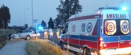Zespół Ratownictwa Medycznego i rozbity samochód osobowy na miejscu wypadku. W samochodzie osobowym uszkodzony przód i bok samochodu. W tle strażacy i radiowóz.