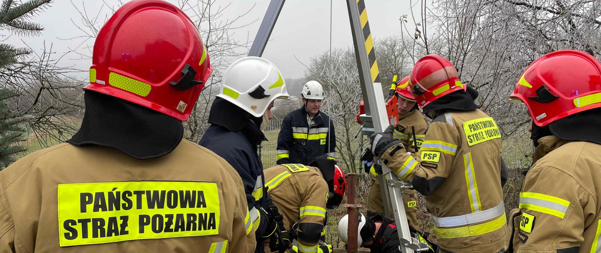 Zdjęcie przedstawia studnię z której strażacy wydobywają ciało kobiety. 
