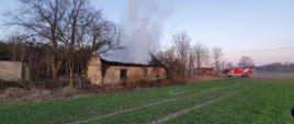 Na zdjęciu widać budynek ze spalonym dachem. Nad budynkiem szary dym. Zdjęcie zrobione w porze porannej. Po prawej stronie samochody ratowniczo-gaśnicze w kolorze czerwonym. Przed budynkiem pole uprawne. Wokół budynku drzewa bez liści. 