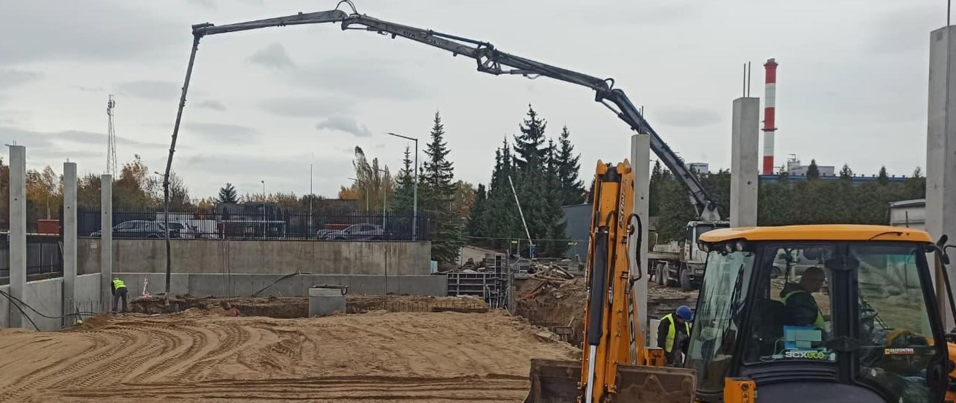 Na zdjęciu plac budowy. Po prawej stronie zdjęcia widoczna żółta koparka. W koparce siedzi mężczyzna, a drugi stopi obok pojazdu. W tle kilku innych mężczyzn, wszyscy w żółtych odblaskowych kamizelkach. 