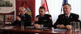 Za stołem prezydialnym komendant wojewódzki i komendant szkoły podoficerskiej PSP w mundurach wyjściowych. Jeden z nich trzyma mikrofon przy ustach. Trzeci, z lewej strony siedzi druh OSP. W tle banery firmowe.