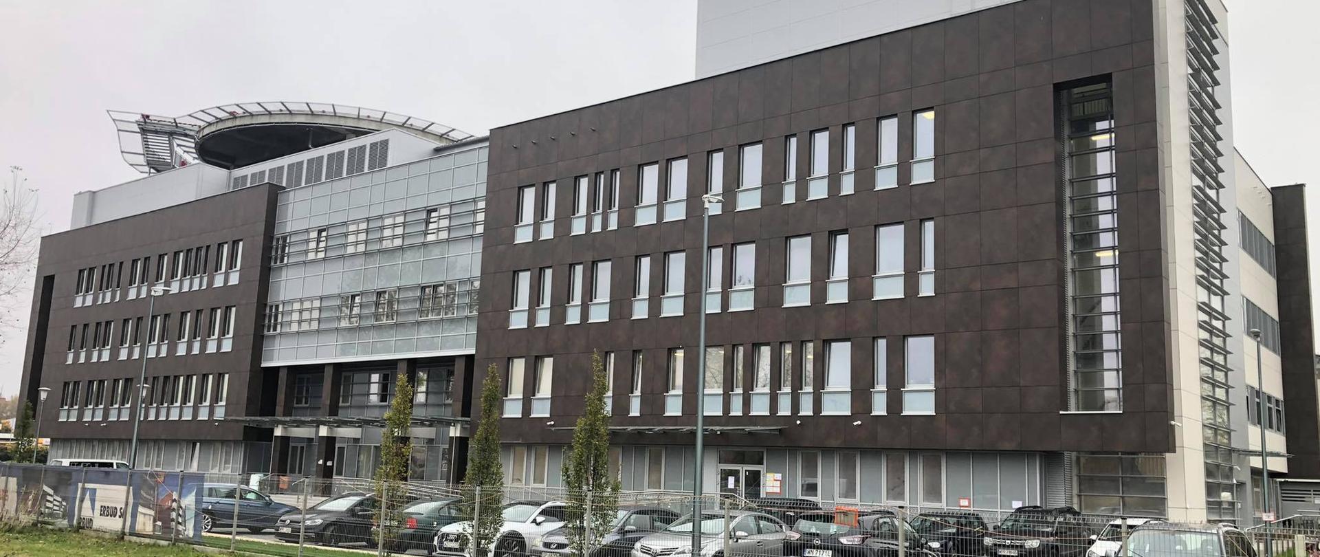 Na zdjęciu znajduje się szaro brązowy budynek szpitalny. Przed budynkiem stoją samochody. Pomiędzy samochodami a pasem zieleni znajduje się płot