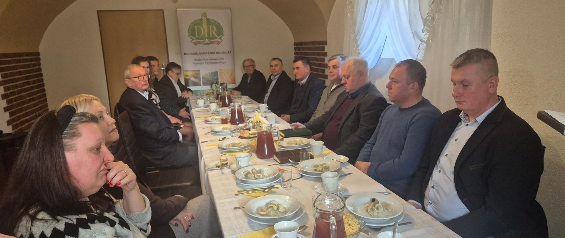 Kilkunastu uczestników spotkania siedzący przy długim stole. W tle znajduje się plakat Dolnośląskiej Izby Rolniczej, a na stole rozstawione są talerze z jedzeniem, w tym pierogi oraz filiżanki i szklanki. W sali panuje przytulna atmosfera, z delikatnym oświetleniem.