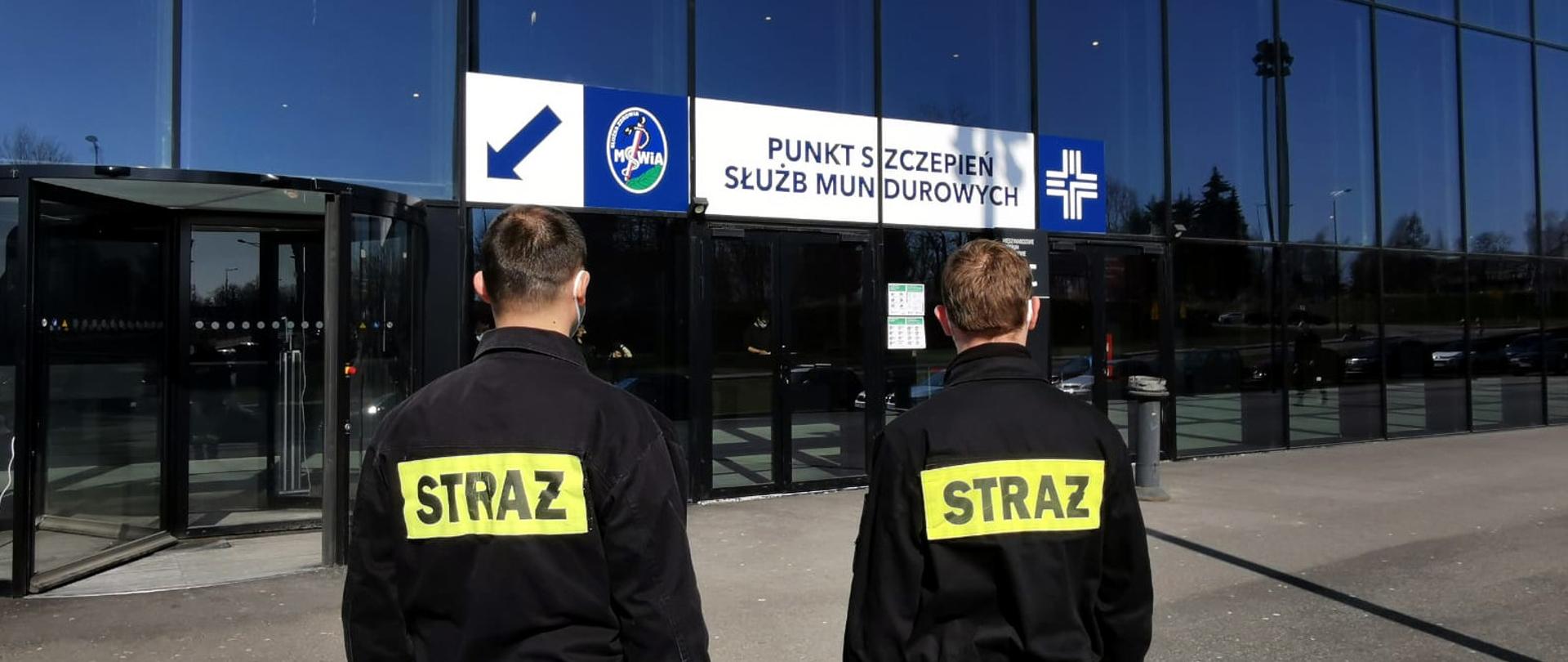 Na zdjęciu widoczny jest budynek Międzynarodowego Centrum Kongresowego przed budynkiem stoi odwróconych tyłem dwóch strażaków w ubraniach koszarowych w oczekiwaniu na szczepienie przeciwko Covid 19.