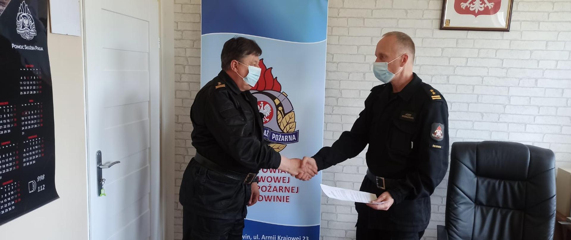 Zdjęcie przedstawia dwóch mężczyzn ubranych w czarne mundury strażackie oraz maseczki chirurgiczne podających sobie ręce, jeden z nich trzyma w rękach dokumenty. Za nimi widnieje baner z logiem PSP oraz napisem: "Komenda Powiatowa Państwowej Straży Pożarnej w Świdwinie". Za banerem widzimy ścianę z tapetą w szare cegły. Na ścianie wisi Godło Rzeczypospolitej Polskiej. Zdjęcie wykonano w gabinecie Komendanta Powiatowego PSP w Świdwinie.