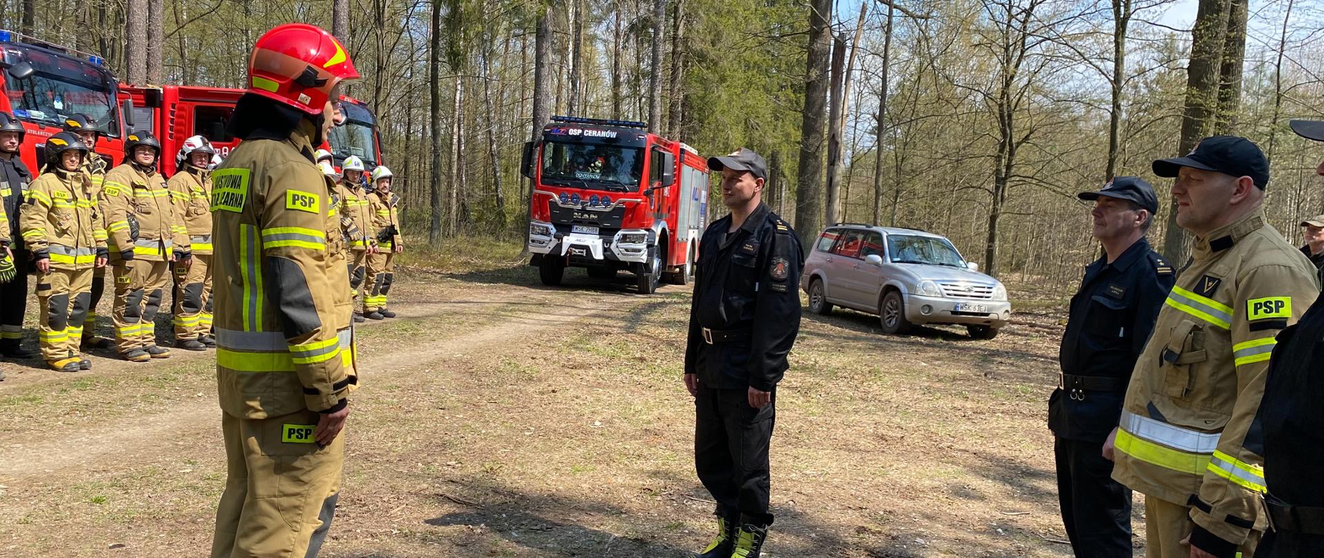 strażacy podczas Ćwiczenia „Kostki 2022”. pododdział druhów stoi na zbiórce przed samochodami pożarniczymi na drodze w lesie. Dowódca melduje Komendantowi zbiórkę.