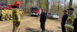 strażacy podczas Ćwiczenia „Kostki 2022”. pododdział druhów stoi na zbiórce przed samochodami pożarniczymi na drodze w lesie. Dowódca melduje Komendantowi zbiórkę.