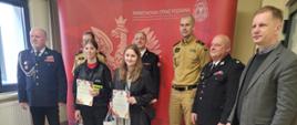 Komendant Miejski w mundurze wyjściowym. Obok niego strażacy w mundurach i pan w marynarce. Dwie dziewczyny trzymające w rękach dyplomy i trorby papierowe stoją przed dorosłymi. Za nimi czerwona ścianka firmowa z dużym orłem białym, napisem Państwowa Straż Pożarna i logo PSP.