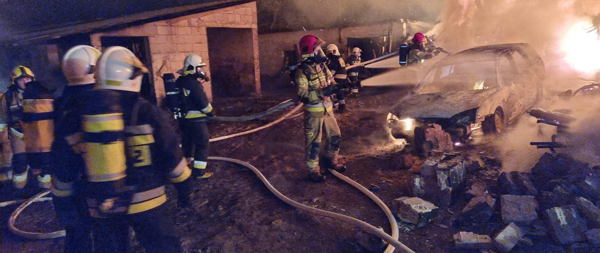 Po prawej stronie gaszony przez strażaków samochód osobowy. W tle ogień. Po środku i po lewej stronie pracujący strażacy